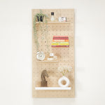 Standard pegboard  - wall organizer with four wooden shelves and white bottom brackets, styled with books, a plant, a camera, an alarm clock, a horse figurine, and a white donut-shaped vase against a white wall.