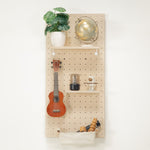 Standard pegboard - wall organizer mounted on a white wall, styled with a potted green plant, a brass globe, a wooden ukulele, small decor items, and a fabric storage pouch at the bottom.