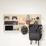 Standard pegboard - wall organizer mounted on a white wall, styled with various tech and travel accessories including a black backpack, bicycle helmet, keyboard, game controllers, sunglasses, and a cap.