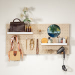 Standard pegboard - wall organizer mounted on a white wall, styled with a brown handbag on a top shelf, a small amber bottle with dried flowers, a round mirror, jewelry hanging from hooks, skincare bottles on a lower shelf, and a black hair dryer hanging on the right.