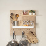 Small pegboard - kitchen wall organizer mounted on a beige wall, holding wooden utensils in a metal cup, spice jars, two small potted succulents, and pots and oven mitts hanging below.