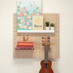 Small pegboard -  wall organizer mounted on a white wall, styled with a colorful floral art print, a book, small potted succulents, a stack of books, a small wooden bird figurine, a brown ukulele hanging from a peg, and a white shelf.