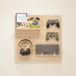 Small pegboard - styled with various tech accessories including black over-ear headphones, a black keyboard, two video game controllers, a green alarm clock, and a small framed picture.