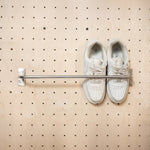 A pair of off-white leather sneakers hanging from a metal rail mounted on a light wood pegboard wall.