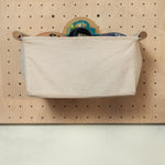 A beige canvas utility basket with wooden handles, hanging on a light wood pegboard wall.