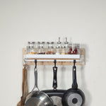 Tiny pegboard - kitchen spice rack with glass jars and bottles on top and three hanging cookware pots below, shown against a plain light wall.