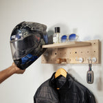 Tiny pegboard - A hand holds a black and blue full-face motorcycle helmet next to a light wood pegboard wall rack. The rack has a shelf holding sunglasses and a small bottle, and hooks holding a black jacket on a wooden hanger and a car key.
