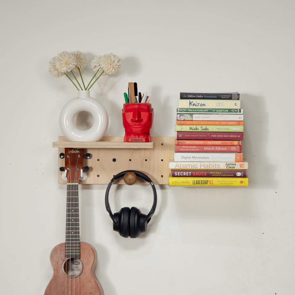 Tiny pegboard -  with pegs and hooks, styled with a white vase of flowers, a red pencil holder, a stack of books, black over-ear headphones, and part of a wooden ukulele against a plain off-white wall.
