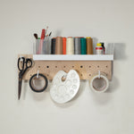Tiny pegboard - with a white shelf and hanging hooks, styled with craft supplies including scissors, thread spools, tape, and an artist palette against a plain wall. 